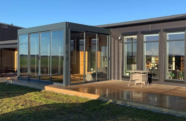 Zimní zahrada v moderní hliníkové konstrukci s velkými posuvnými skleněnými plochami plynule navazuje na dřevěnou terasu a obývací část rodinného domu. Díky celoproskleným stěnám nabízí celoročně světlý prostor s výhledem do otevřené krajiny.